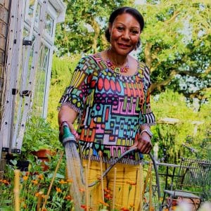 A portrait photo of Andrea in a garden, wearing iconic colours, smiling at the camera and watering plants in preparation for her Gardener's World exhibition.