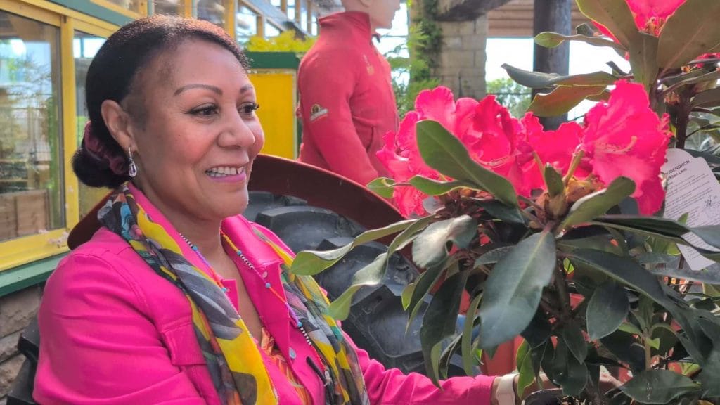 A close up of Andrea holding a plant, which has vibrant pink flowers that match her jacket perfectly.